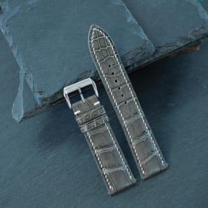 May include: A grey watch strap featuring a textured alligator-style embossed pattern with white contrast stitching. The strap includes a polished silver-tone metal buckle and two matching leather keepers, designed for secure attachment to a timepiece.