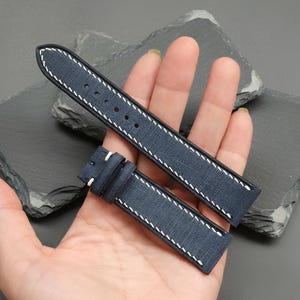 May include: A navy blue watch strap with white stitching. The strap is made of a textured material and has a buckle. The strap is held in a hand.