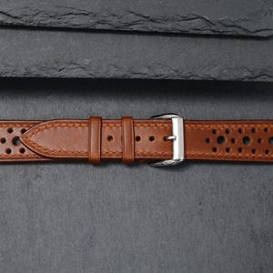 K&ouml;nnte beinhalten: Ein braunes Leder-Uhrenarmband mit perforiertem Design. Das Armband hat eine silberne Schnalle und ist mit einem helleren braunen Faden gen&auml;ht. Das Uhrenarmband wird auf einer dunkelgrauen Oberfl&auml;che pr&auml;sentiert.