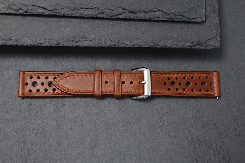 K&ouml;nnte beinhalten: Ein braunes Leder-Uhrenarmband mit perforiertem Design. Das Armband hat eine silberne Schnalle und ist mit einem helleren braunen Faden gen&auml;ht. Das Uhrenarmband wird auf einer dunkelgrauen Oberfl&auml;che pr&auml;sentiert.