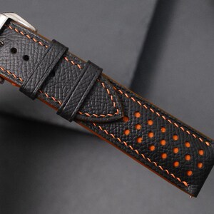 May include: Close-up of a black leather watch strap with orange stitching and a silver buckle. The strap has a textured surface and orange perforations along the edge. The stitching is a contrasting orange colour.
