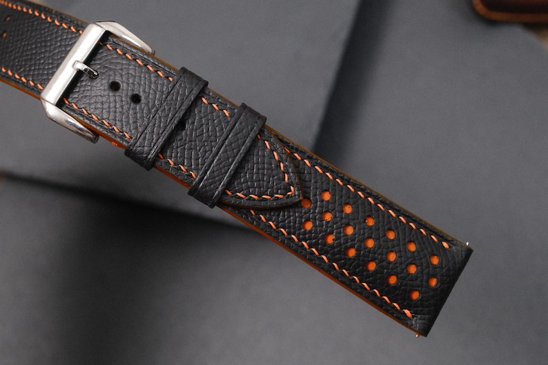 May include: Close-up of a black leather watch strap with orange stitching and a silver buckle. The strap has a textured surface and orange perforations along the edge. The stitching is a contrasting orange colour.