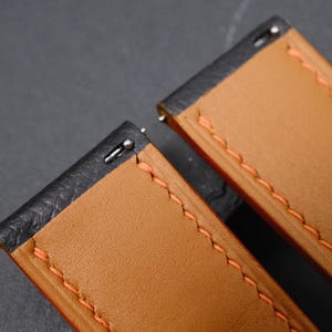 May include: Close-up of two watch straps. The straps are made of tan leather with orange stitching. The edges are black. The metal pin is visible at the end of each strap.