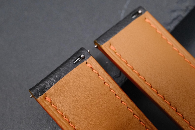 May include: Close-up of two watch straps. The straps are made of tan leather with orange stitching. The edges are black. The metal pin is visible at the end of each strap.