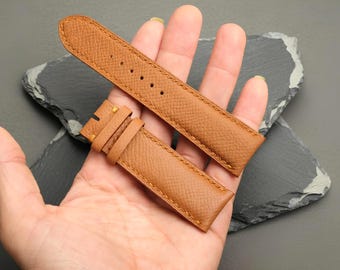 May include: A tan leather watch strap featuring a textured cross-hatch pattern, tonal stitching along the edges, and a series of punched holes for adjustment. The strap consists of two separate pieces, one with a buckle loop and one with holes.