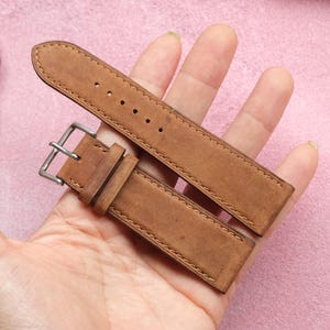 Puede incluir: Una correa de reloj de cuero marrón claro con una hebilla plateada. La correa está hecha de un material de cuero texturizado y tiene costuras visibles a lo largo de los bordes. La correa del reloj se muestra sobre un fondo rosa.