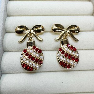 May include: A pair of gold-tone drop earrings featuring a bow at the top and a round ornament pendant below. The ornament is decorated with diagonal stripes of red rhinestones and small white faux pearls, creating a festive, holiday-inspired design.