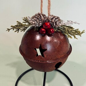May include: A rustic, brown metal Christmas bell ornament with a star cutout and a small hole. The ornament is decorated with faux pinecones, red berries, and greenery, and hangs from a twine loop. The bell is supported by a black metal stand.