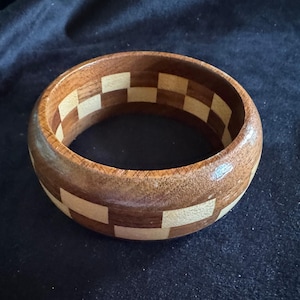 May include: A wooden bangle bracelet with a checkerboard pattern. The bracelet is made of alternating light and dark brown wood squares. The bracelet is a wide, circular shape.