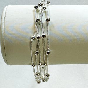May include: A multi-strand silver bracelet featuring thin, flexible snake chains adorned with small, polished silver beads spaced at intervals along each strand. The bracelet is wrapped around a cylindrical display form, showcasing its layered design.