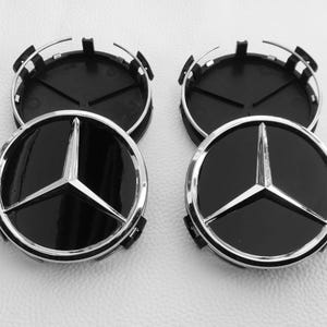 Puede incluir: Cuatro tapacubos negros y plateados con el logotipo de Mercedes-Benz. Los tapacubos tienen un acabado negro brillante con un emblema de estrella plateada de tres puntas. Fondo gris claro.