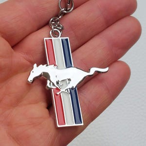 May include: A silver-coloured keyring featuring a running horse emblem. The emblem is set against a vertical bar with red, white, and blue stripes. The keyring is attached to a silver chain.