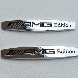 Può includere: Due emblemi cromati AMG Edition. Ogni emblema presenta il logo AMG e la parola "Edition" in testo nero. Gli emblemi rettangolari hanno bordi arrotondati e sono esposti su una superficie bianca.