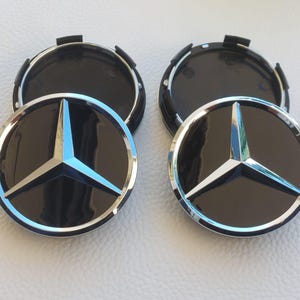 Op de afbeelding: Twee zwarte en zilveren Mercedes-Benz wieldopjes. Elke dop heeft een zilveren sterembleem met een lichtblauw centrum op een zwarte achtergrond, omgeven door een zilveren ring.