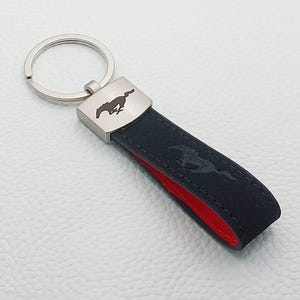 Puede incluir: Llavero de cuero negro con un acento metálico plateado con un logotipo de caballo. El llavero tiene un interior rojo y un llavero plateado. El logotipo del caballo también está grabado en la correa de cuero negro.