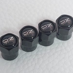 Peut inclure: Quatre bouchons de valve de pneu noirs, hexagonaux, avec le logo "OZ Racing" argenté. Ces bouchons brillants sont conçus pour les passionnés d'automobile.