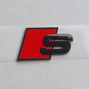 Puede incluir: Un emblema S negro y rojo sobre un fondo blanco. El emblema tiene un acabado brillante y probablemente es para un vehículo. La sección roja tiene forma de trapecio y la "S" negra está estilizada.