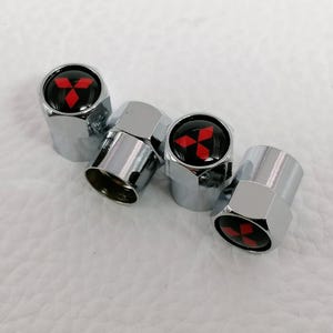 Peut inclure: Quatre bouchons de valve chromés. Chaque bouchon a un dessus noir avec un logo rouge à trois diamants. Les bouchons sont hexagonaux et ont une surface métallique brillante et réfléchissante.