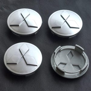 Puede incluir: Cuatro tapacubos plateados Mitsubishi. Cada tapa presenta el logotipo de Mitsubishi, un diseño de tres diamantes. Una tapa está separada, revelando la parte trasera de plástico gris. Las tapas están sobre una superficie oscura.