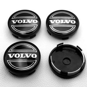 Puede incluir: Cuatro tapacubos negros de Volvo, cada uno con el nombre de la marca en letras blancas. Los tapacubos son redondos con un contorno plateado y un centro negro. Se muestra un tapacubos desde atrás.