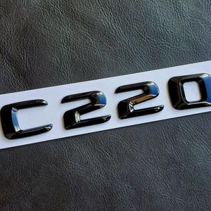 May include: Glossy black C220 emblem on a white strip against a textured dark background. The emblem features a bold, modern font, with each character clearly defined and reflective.