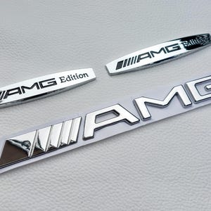 Op de afbeelding: Drie chromen AMG-emblemen op een witte ondergrond. Twee kleinere emblemen lezen "AMG Edition". Het grootste embleem spelt "AMG" in afzonderlijke letters. Alle emblemen hebben een glanzende, reflecterende afwerking.
