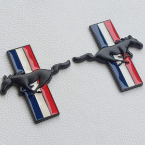 May include: Two black Mustang emblems. Each emblem features a running horse over a rectangular shape with red, white, and blue stripes. The emblems are set against a white textured background.
