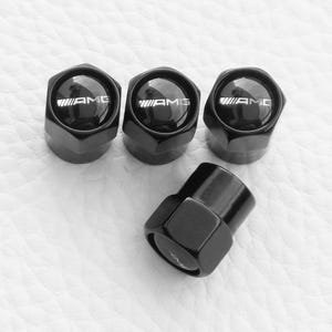 Peut inclure: Bouchons de valve de pneu AMG noirs. Les bouchons sont hexagonaux et arborent le logo AMG en blanc. Ils sont disposés sur une surface blanche texturée.