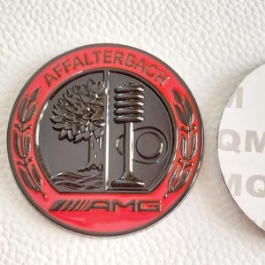 Puede incluir: Un emblema redondo, negro y rojo con el texto "AFFALTERBACH" y "AMG". El emblema presenta un árbol y un diseño estilizado. Un respaldo adhesivo blanco es visible a la derecha.