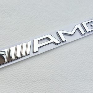 Può includere: Un emblema AMG cromato su un supporto bianco. L'emblema presenta le lettere "AMG" in un carattere stilizzato, con un caratteristico design a tre barre sul lato sinistro. L'emblema è progettato per applicazioni automobilistiche.