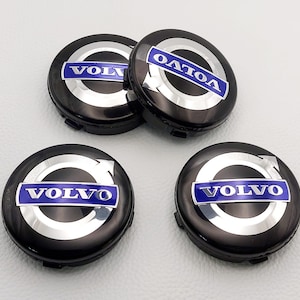 Puede incluir: Cuatro tapacubos Volvo negros con un anillo plateado y texto azul. La palabra "VOLVO" se muestra en letras blancas sobre un fondo azul. Las tapas son redondas y tienen un acabado brillante.