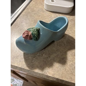 Può includere: Un vaso o contenitore decorativo in ceramica a forma di zoccolo azzurro, caratterizzato da una rosa rosa scolpita e foglie verdi applicate sulla parte superiore. L'oggetto presenta una finitura liscia e lucida con un interno cavo.