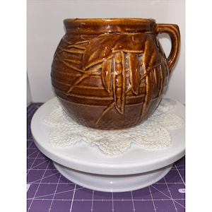May include: A brown ceramic mug with a handle and a textured surface. The mug features a relief design of leaves and pods. The mug is sitting on a white, tiered cake stand with a lace doily.