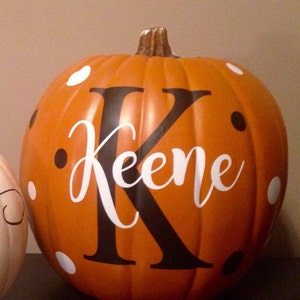 May include: An orange pumpkin with a black letter K and the name Keene written in white cursive on the front. The pumpkin is decorated with white and black polka dots.