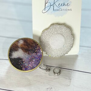 May include: Two round, gold-rimmed, glitter-covered badge reels. One reel has a brown and white cowhide pattern with purple glitter. The other reel has a white glitter design. The reels are on a white wooden surface. B Keene Kreations is written on a white card behind the reels.