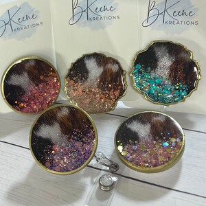 May include: Four round, resin-coated, cowhide-patterned, glitter-filled keychains with gold-colored metal rims. One keychain is attached to a clear plastic retractable badge holder with a silver-colored metal clip.