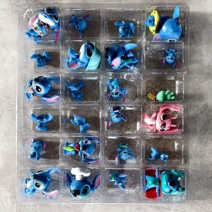 K&ouml;nnte beinhalten: Eine transparente Kunststoffschale gef&uuml;llt mit kleinen, blauen Stitch-Figuren in verschiedenen Posen. Einige Figuren haben Accessoires wie Sonnenbrillen oder Kochm&uuml;tzen. Eine rosa Figur ist ebenfalls vorhanden. Die Figuren befinden sich in einzelnen F&auml;chern.