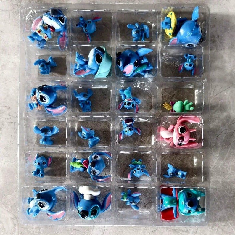 K&ouml;nnte beinhalten: Eine transparente Kunststoffschale gef&uuml;llt mit kleinen, blauen Stitch-Figuren in verschiedenen Posen. Einige Figuren haben Accessoires wie Sonnenbrillen oder Kochm&uuml;tzen. Eine rosa Figur ist ebenfalls vorhanden. Die Figuren befinden sich in einzelnen F&auml;chern.