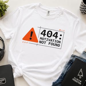 Peut inclure: Un t-shirt blanc à manches courtes avec une impression graphique représentant un triangle orange avec un point d'exclamation noir à côté du texte 404: MOTIVATION NOT FOUND, encadré par un fin rectangle noir avec des icônes géométriques.