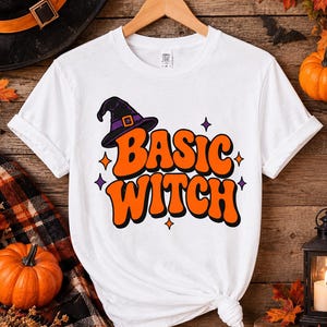 Op de afbeelding: Een wit t-shirt met korte mouwen en een grafische print met de tekst "BASIC WITCH" in dikke oranje letters met een zwarte omlijning. Boven de tekst staat een paarse en zwarte heksenhoed, omringd door kleine oranje en paarse vierpuntige sterren.