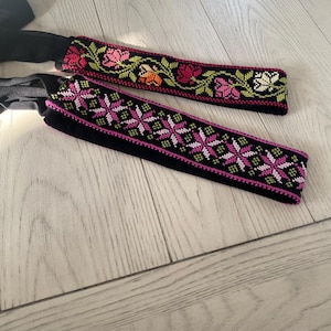 May include: Two black headbands with embroidered floral and geometric patterns. One headband features pink, red, and orange flowers with green leaves. The other has a geometric design in pink and white on a black background.