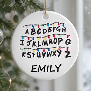 May include: A white ceramic Christmas ornament with a gold string. The ornament features the alphabet with colorful Christmas lights above each letter. The name "EMILY" is written in black below the alphabet.