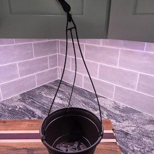 May include: A black hanging planter featuring a round basin with a drainage insert at the bottom. The planter is suspended by three thin, twisted black cords that connect to a single black plastic hook at the top for easy overhead mounting.
