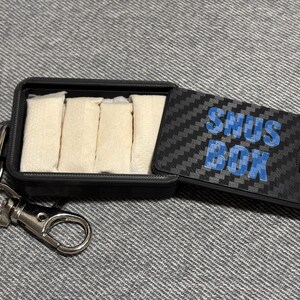 Könnte beinhalten: Eine schwarze Snus-Box mit einem Karbonfasermuster auf dem Deckel und den Worten "SNUS BOX" in Blau. Die Box ist geöffnet und zeigt mehrere weiße Beutel. Ein silberner Schlüsselanhänger ist befestigt.