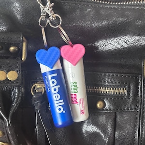 Pode incluir: Dois protetores labiais com chaveiros em forma de coração presos a uma bolsa de couro preta. Um é azul com o texto "Labello Original", o outro é branco com o texto "Spha med". A bolsa tem ferragens douradas.