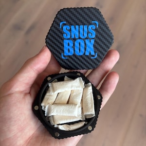 Può includere: Un contenitore esagonale nero con un motivo in fibra di carbonio e il testo blu "SNUS BOX" sul coperchio. Il contenitore è aperto, rivelando diverse bustine bianche all'interno. Il contenitore è tenuto in una mano.