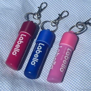 Könnte beinhalten: Drei Labello Lippenbalsam-Tuben in Pink, Blau und Magenta, jeweils mit einem silbernen Schlüsselanhänger. Die Tuben tragen den weißen Schriftzug "Labello" sowie "Velvet Rose" oder "Original". Der Hintergrund ist ein hellblau-weißes Vichy-Muster.