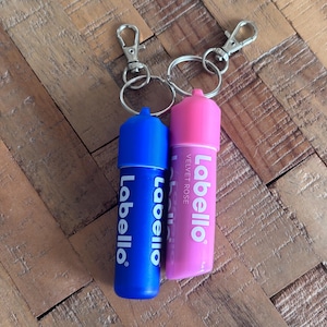Puede incluir: Dos tubos de bálsamo labial Labello, uno azul y otro rosa, sujetos a llaveros. El tubo azul tiene texto blanco, y el tubo rosa dice "Velvet Rose". Ambos tubos tienen llaveros y cierres plateados.