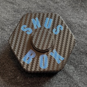 May include: A hexagonal fidget spinner with a carbon fibre pattern and the text "SNUS BOX" in light blue. The spinner has a central circular component and a textured surface.