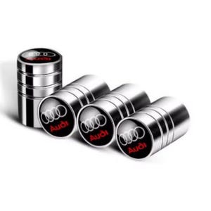 May include: Set of four silver-colored tire valve stem caps with the Audi logo in black and red. One cap is standing upright, while the other three are lying on their sides. The caps have a shiny, metallic finish.